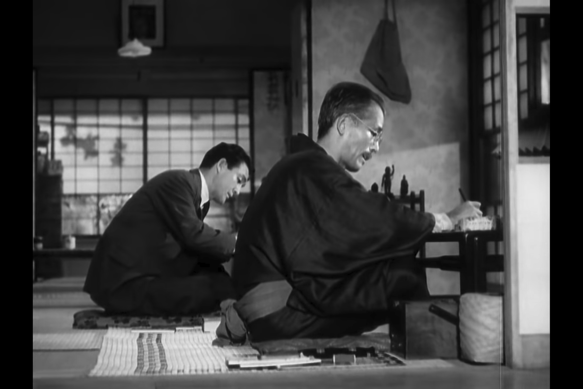 The Weight of Tea: Professor Somiya, wearing a traditional kimono, sits on tatami mats working on Western economics with his assistant Hattori, who wears a Western suit.